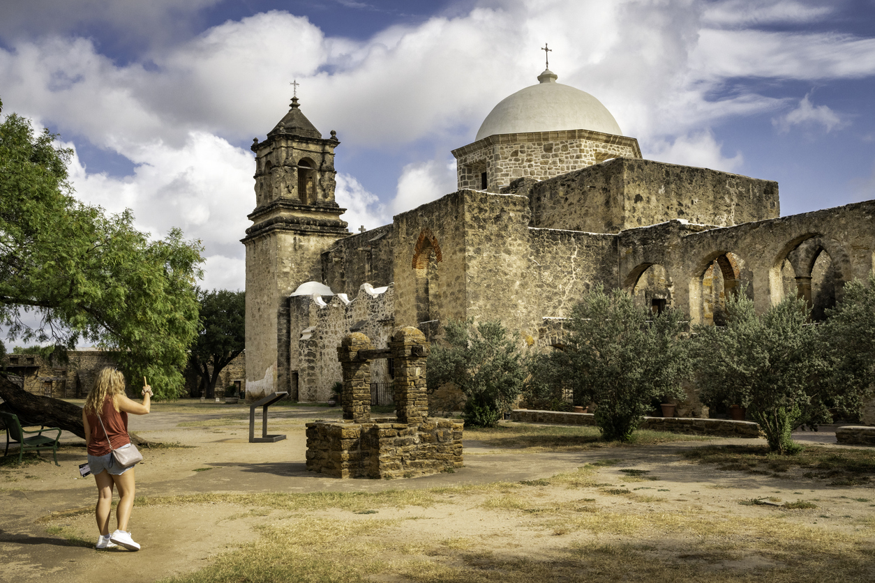 A woman in a red tank top stands in a dirt courtyard taking a photo of a historic stone mission with a bell tower and a domed roof.