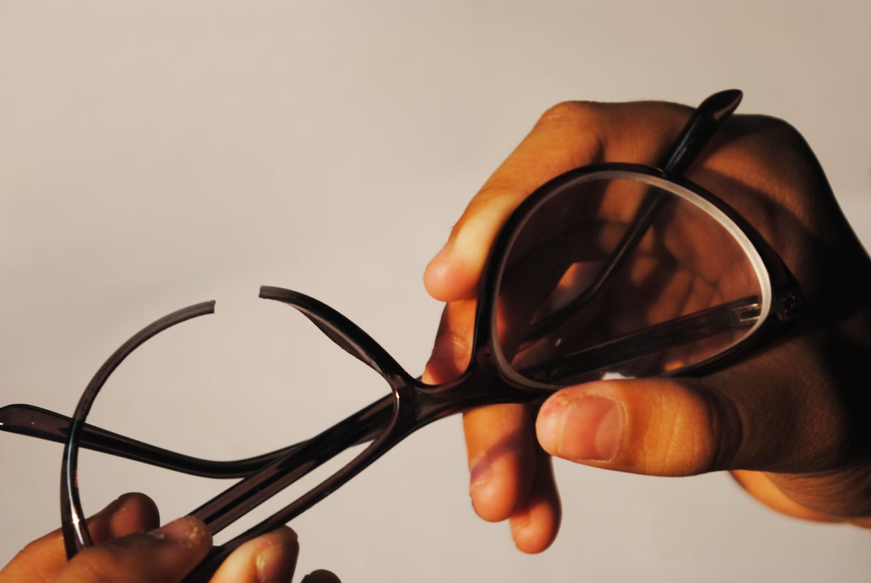 A pair of dark-rimmed glasses with a broken frame being held by two hands.