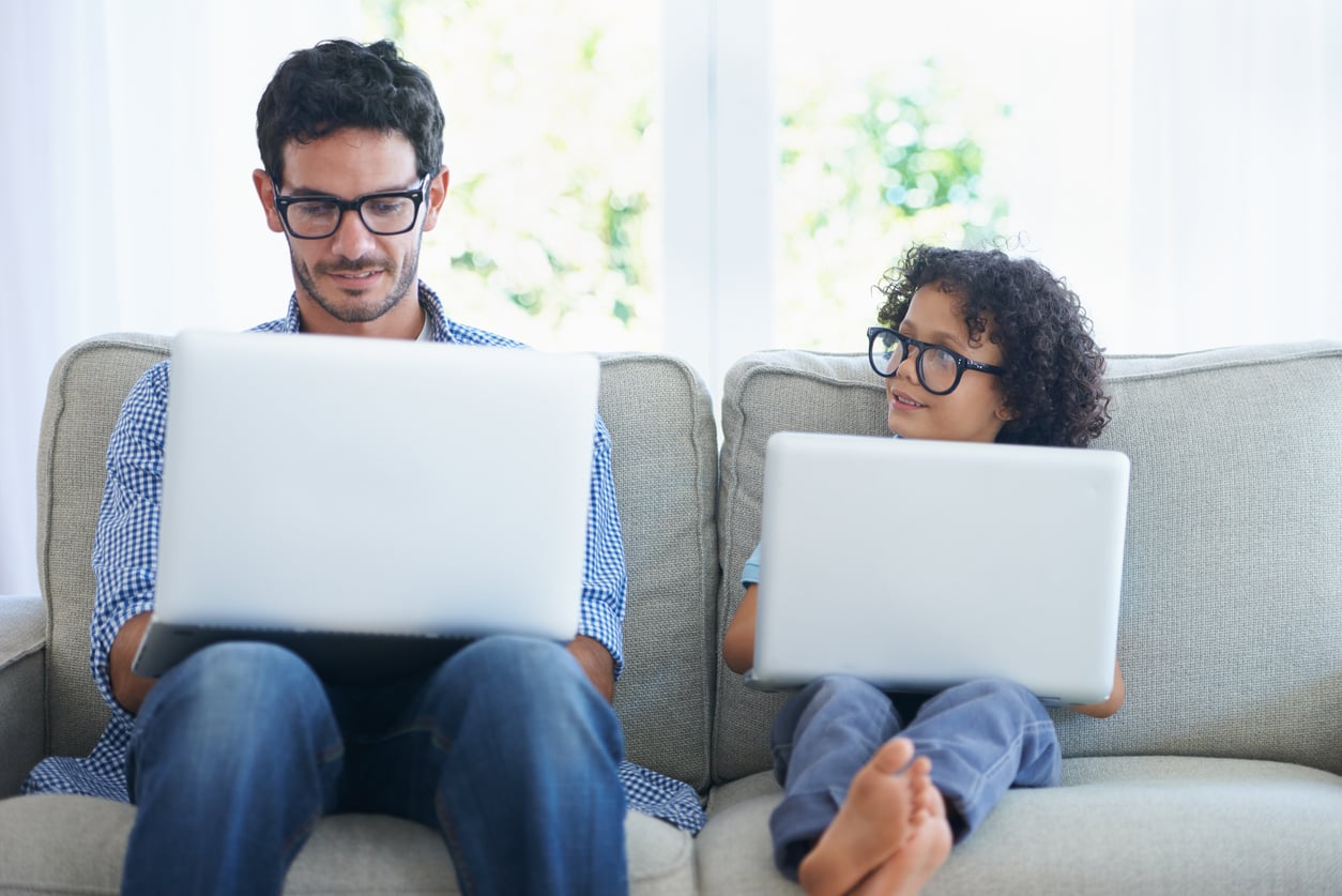 A father and child sitting on a couch, both wearing glasses and using laptops.