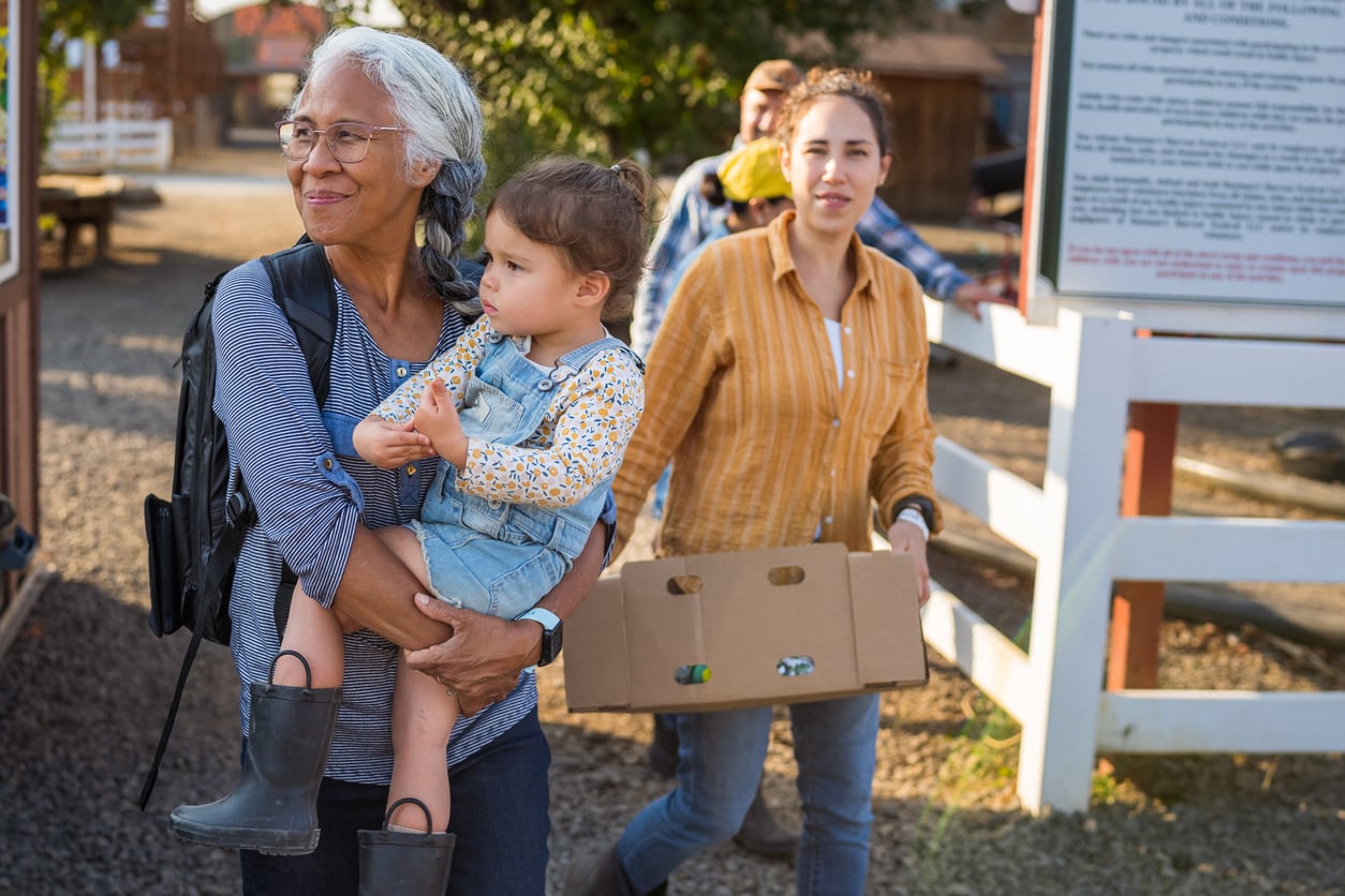 A woman carrying a toddler in denim overalls walks past a woman holding a cardboard box.