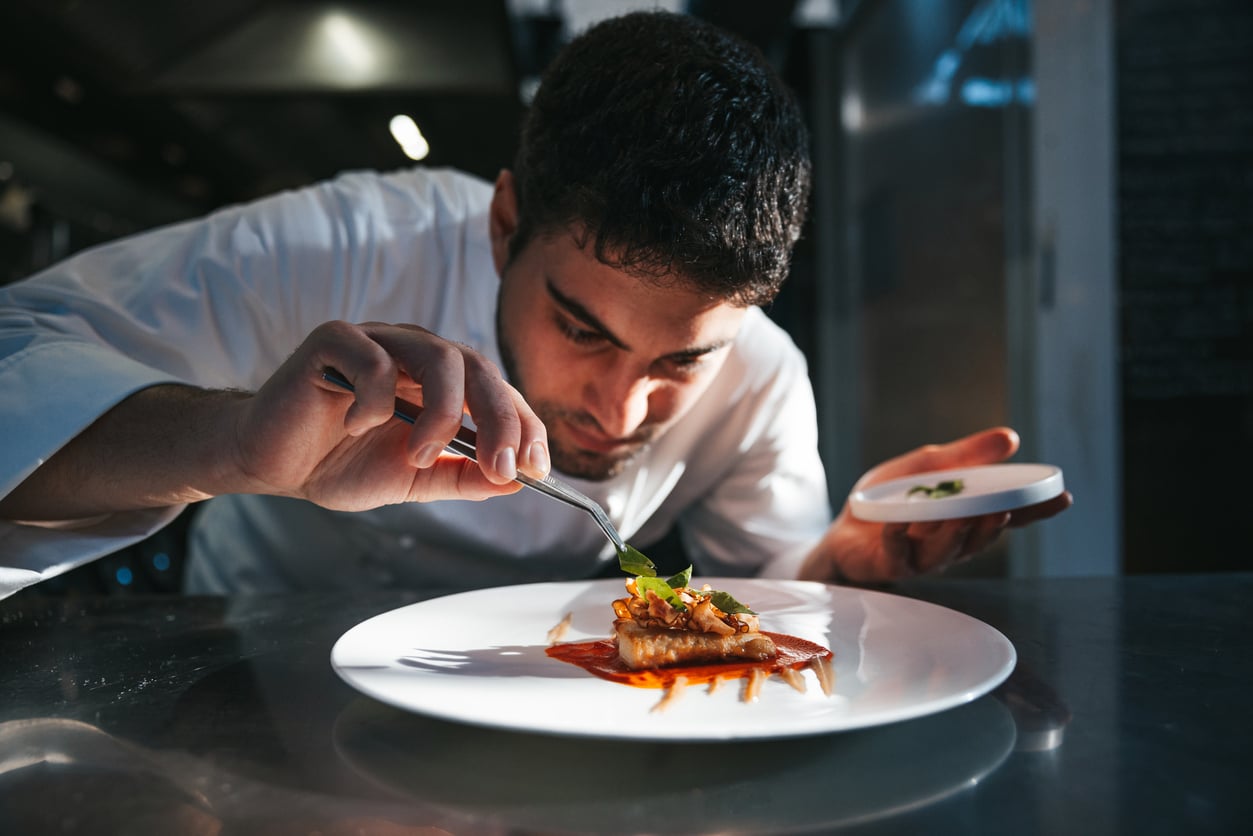 A chef uses tweezers to carefully garnish a plated dish with herbs in a professional kitchen.