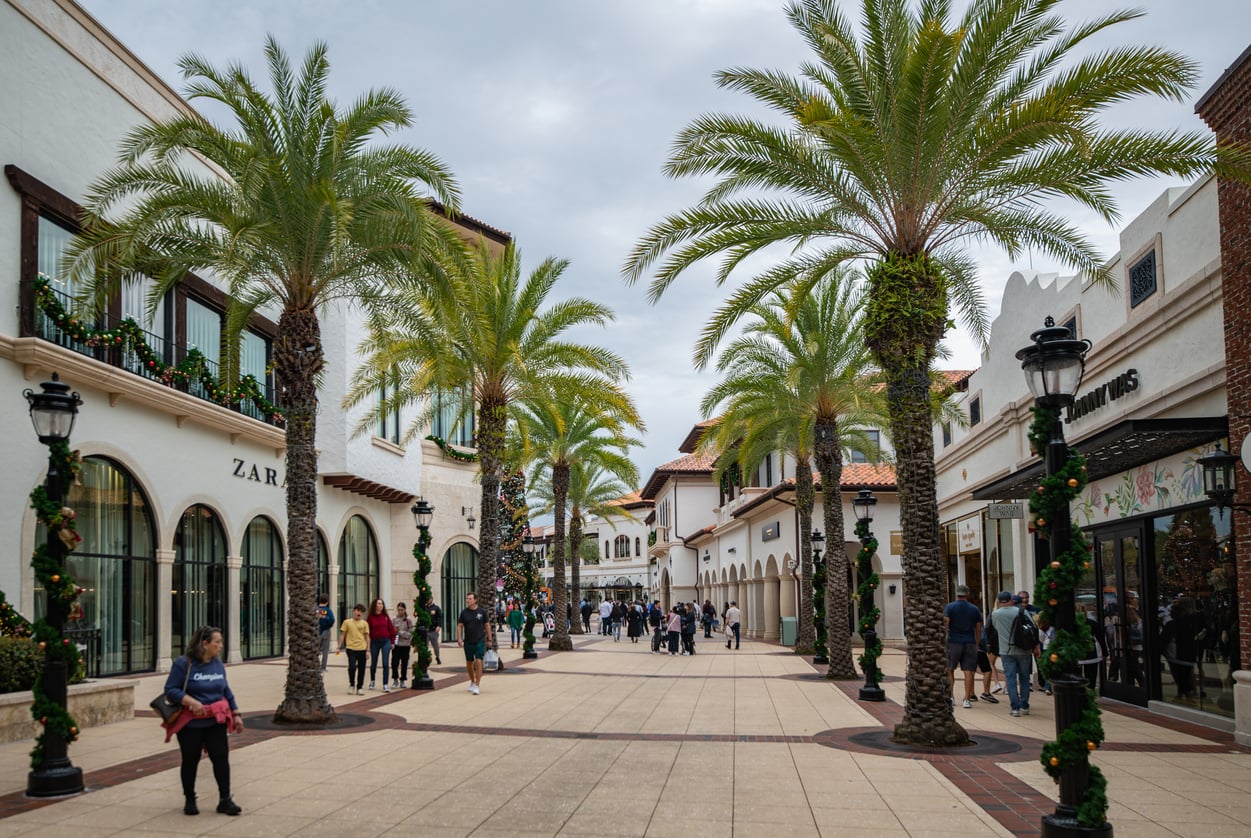 Pedestrians walk along an outdoor shopping promenade lined with palm trees and retail storefronts under a cloudy sky.