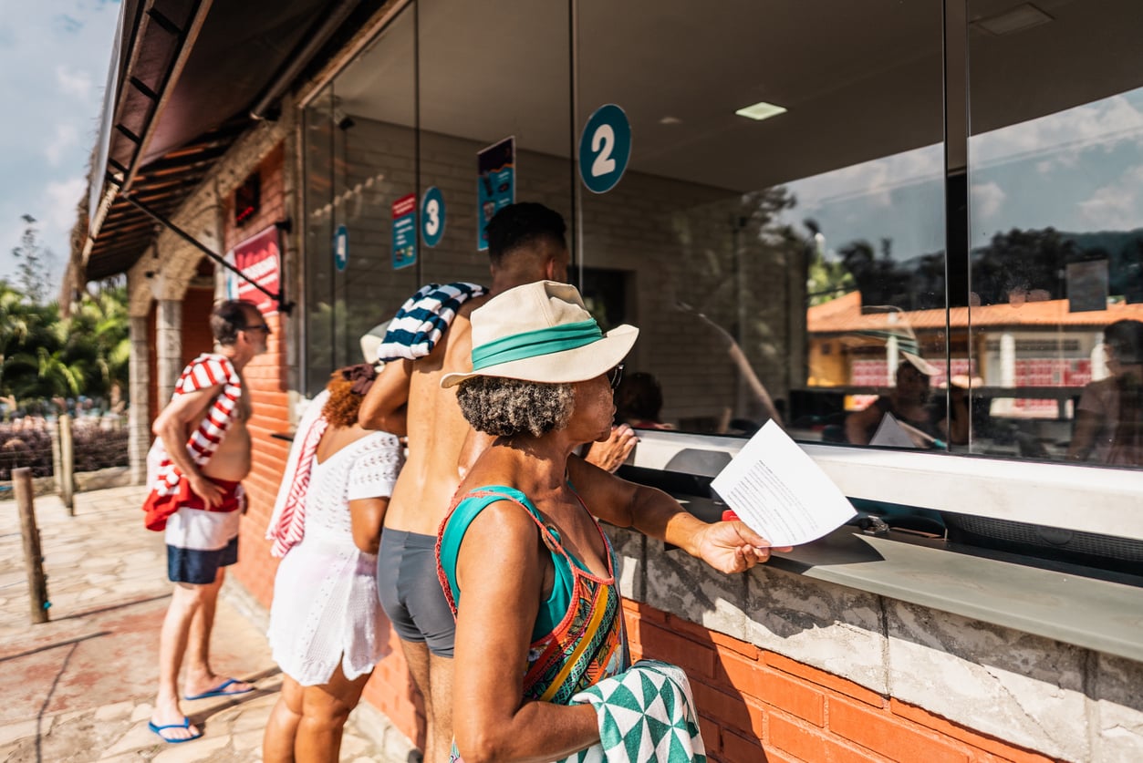 People in swimsuits stand in a line at a ticket window, with one woman handing a paper to the clerk inside.