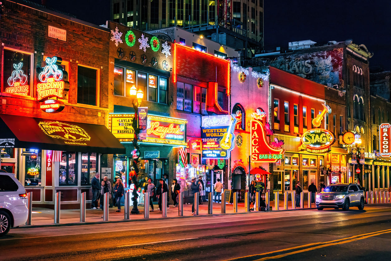 Nighttime street view of Broadway in Nashville, featuring several honky-tonks with bright neon signs, storefronts, and pedestrians walking along the sidewalk.