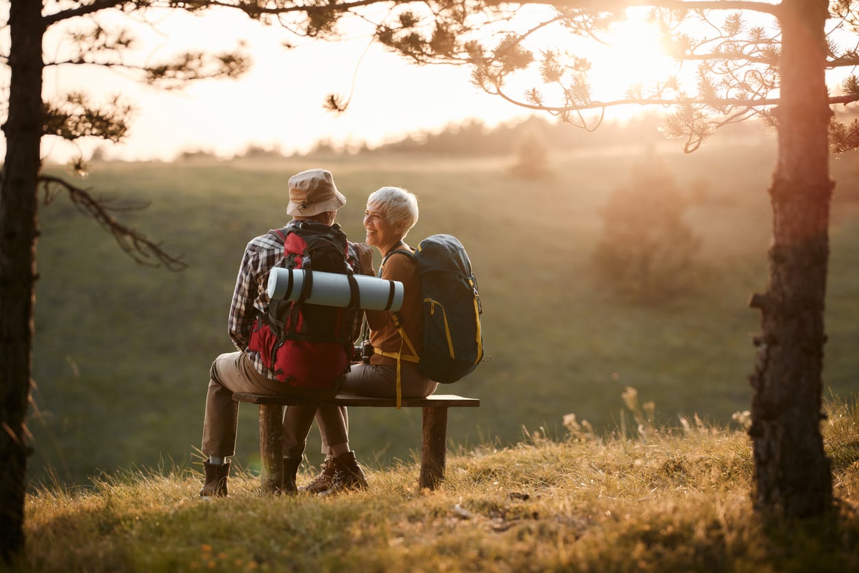 A senior couple with backpacks sits on a wooden bench outdoors at sunset while making trip planning decisions.