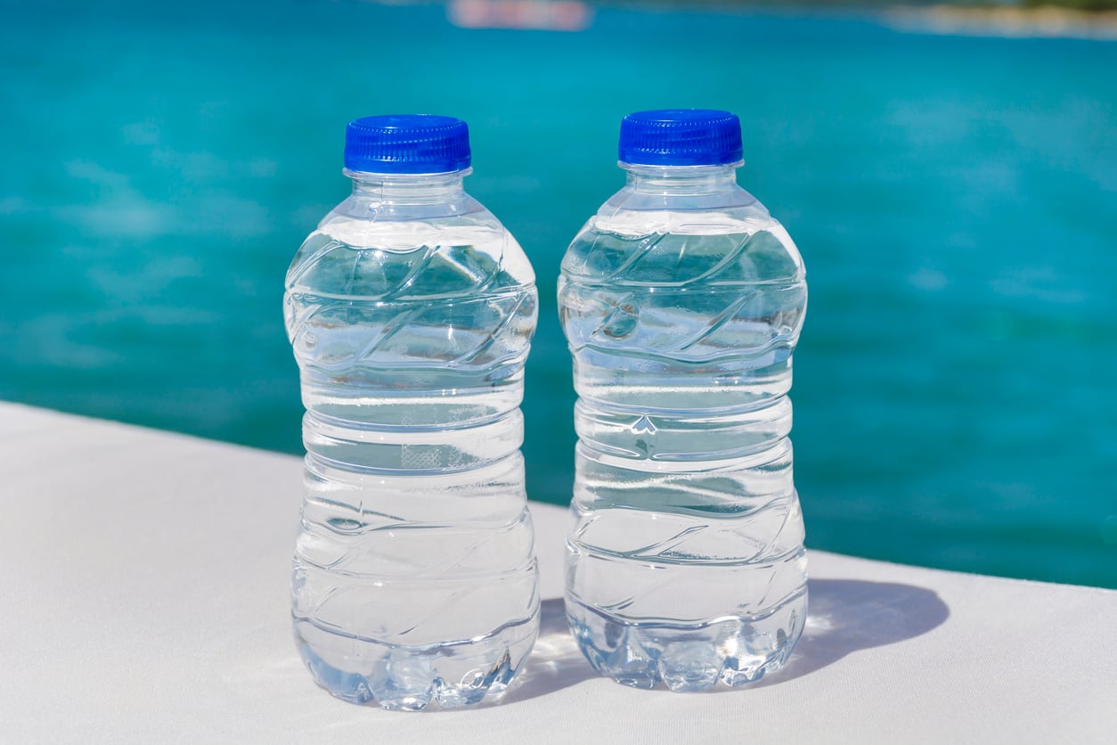 Two plastic water bottles with blue caps sitting on a white surface in front of a blue ocean.