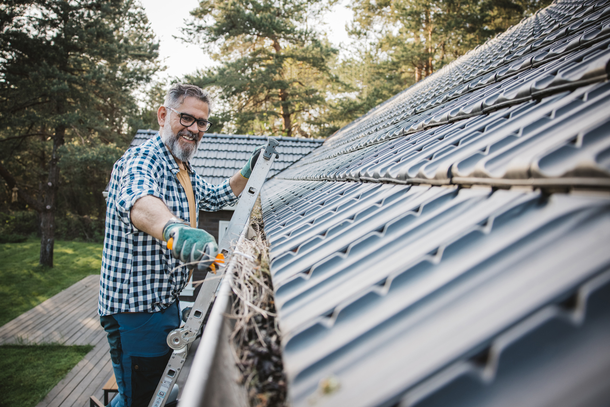 A smiling man on a ladder cleans leaves and debris out of a roof gutter.