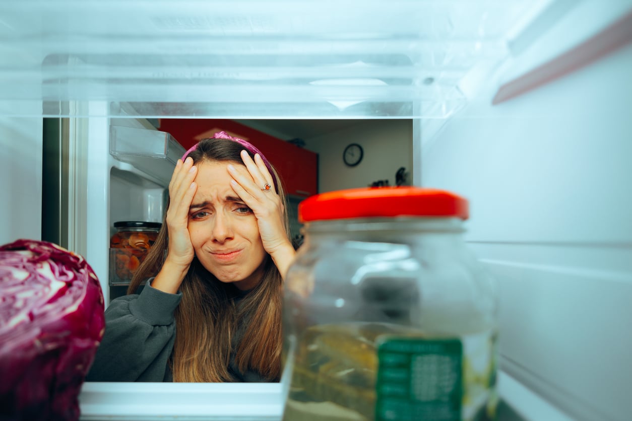 A woman looks into an empty refrigerator with a stressed, frustrated expression, holding her head with both hands.