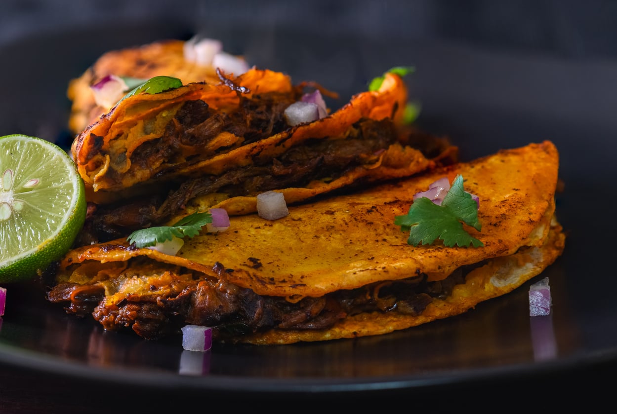 Two beef birria tacos topped with cilantro and onion on a black plate, served with a lime wedge.