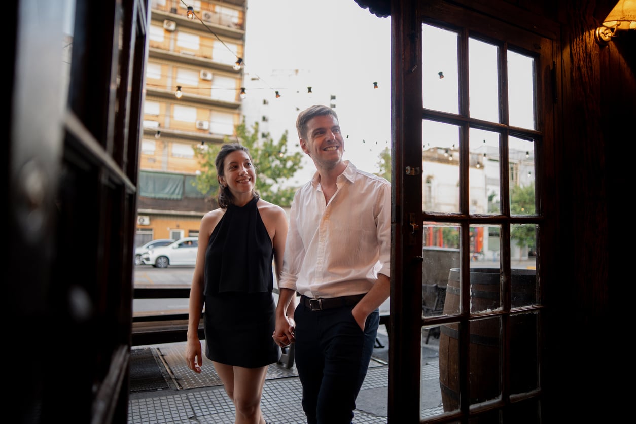 A couple holding hands walks through an open doorway onto a city sidewalk.