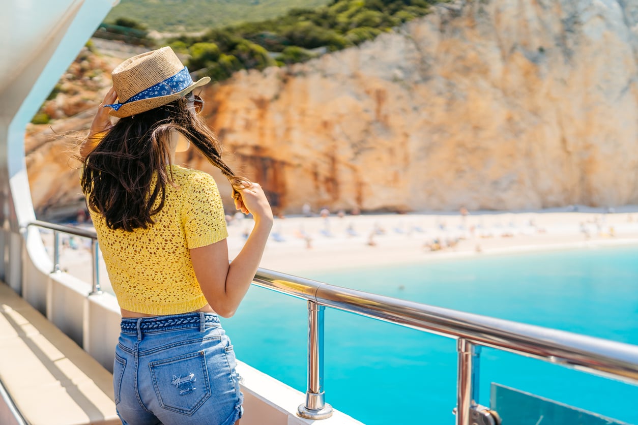 A person in a straw hat and yellow top stands on a boat deck, looking out at a turquoise bay and a sandy beach framed by cliffs.