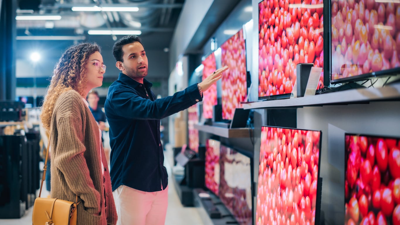 A man points to a television screen displaying a pattern of red tomatoes while a woman looks on in an electronics store.