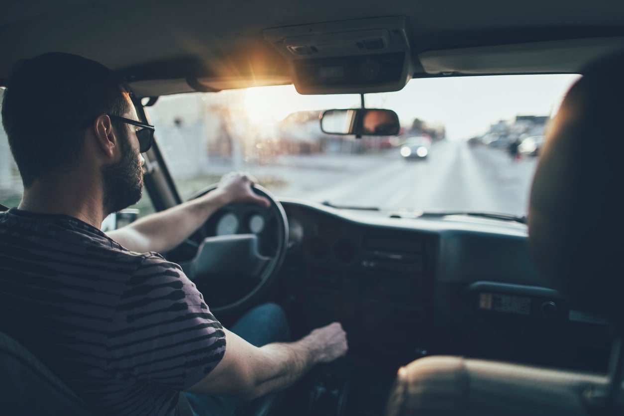A man wearing sunglasses drives a car on a sunlit road.