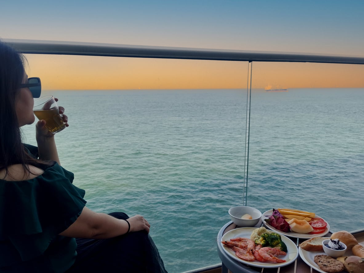 A woman wearing sunglasses drinks from a glass on a balcony table set with plates of shrimp, fruit, and bread overlooking the ocean at sunset, avoiding any hidden cruise ship charges.