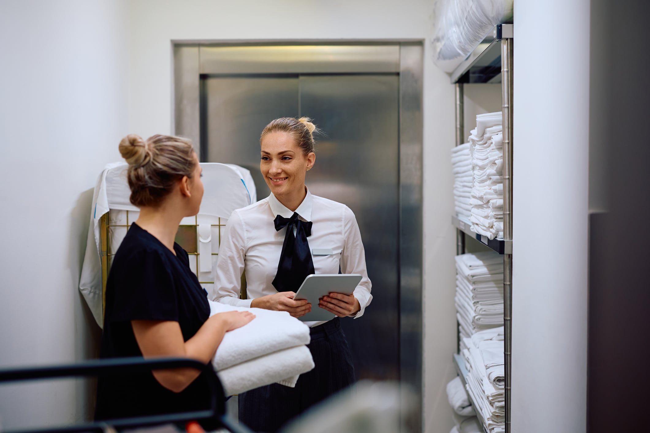 Two hotel staff members in uniform talk in a supply closet, one holding a tablet and the other holding a stack of clean towels.