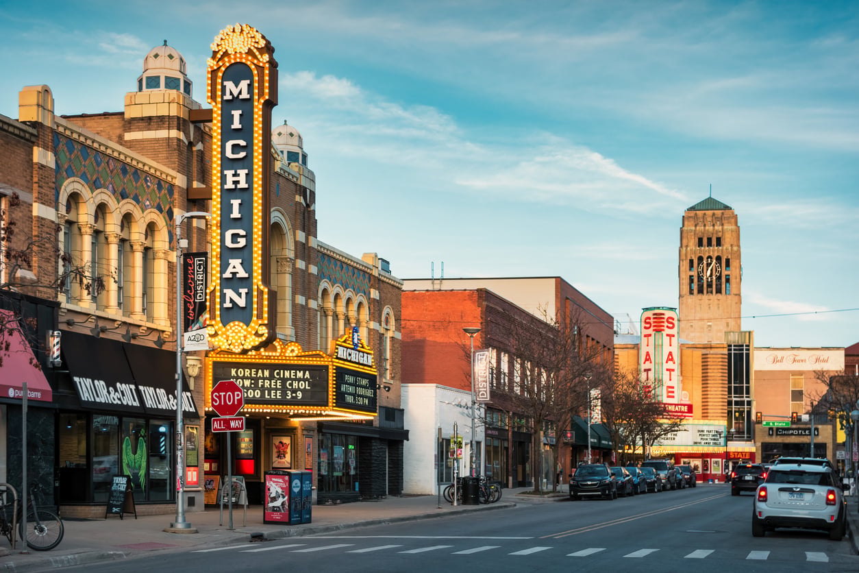 Street view of downtown Ann Arbor, Michigan, featuring the brightly lit marquee of the Michigan Theater and the distant Burton Memorial Tower.