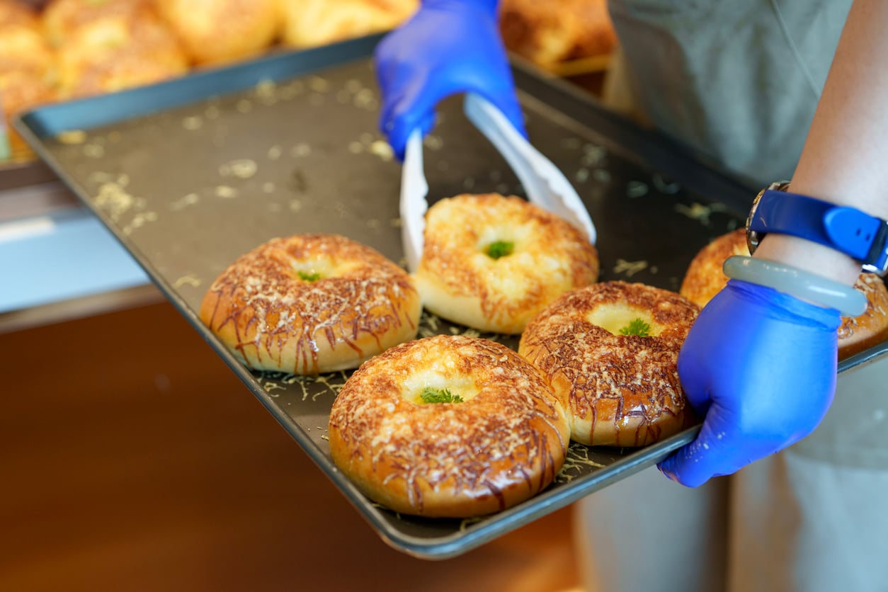 A person wearing blue gloves uses tongs to place a cheese-topped roll onto a baking tray filled with other rolls.