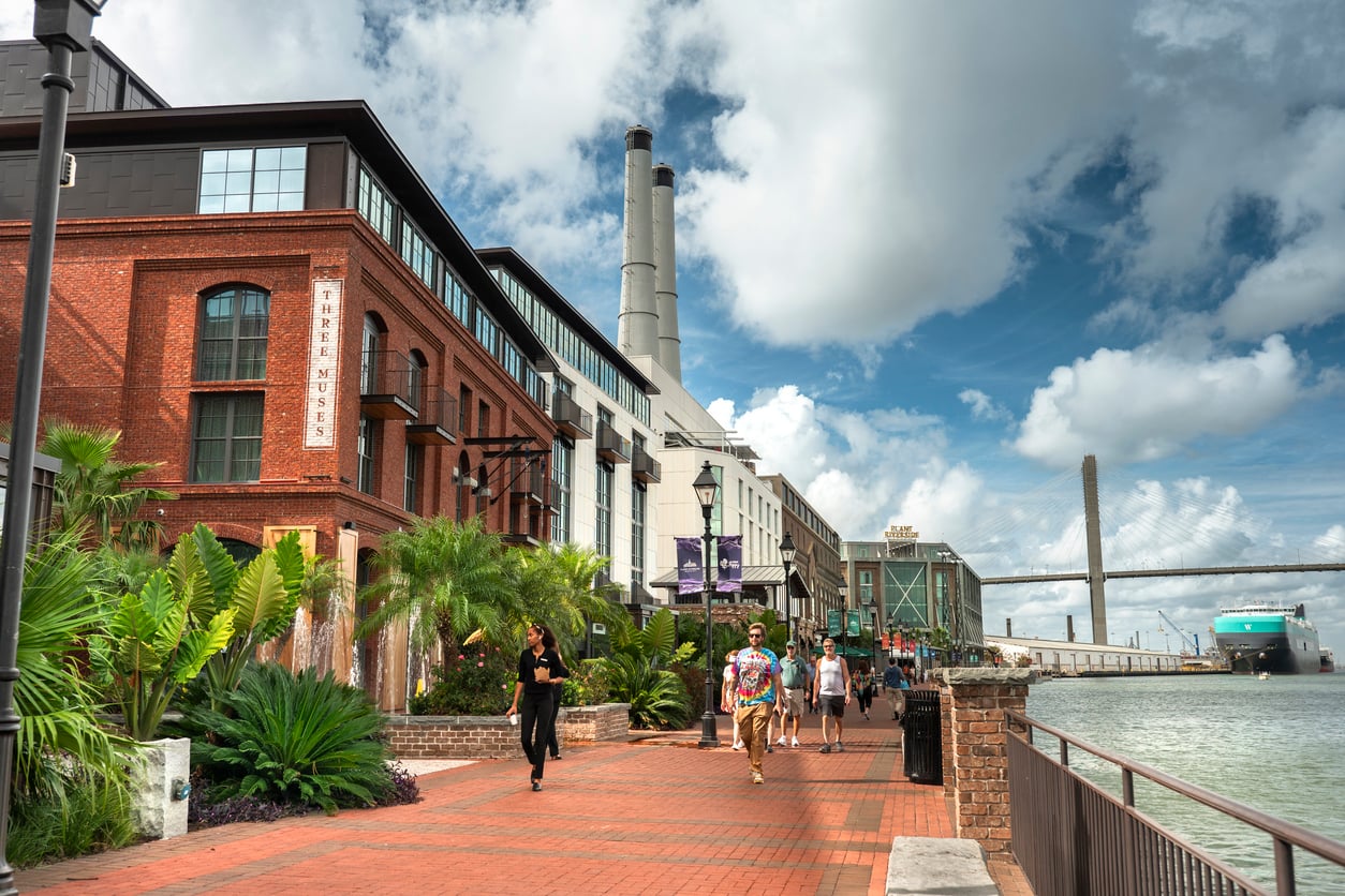 People walking along a brick promenade beside a river in Savannah, Georgia, with the Talmadge Memorial Bridge and a large cargo ship in the distance.