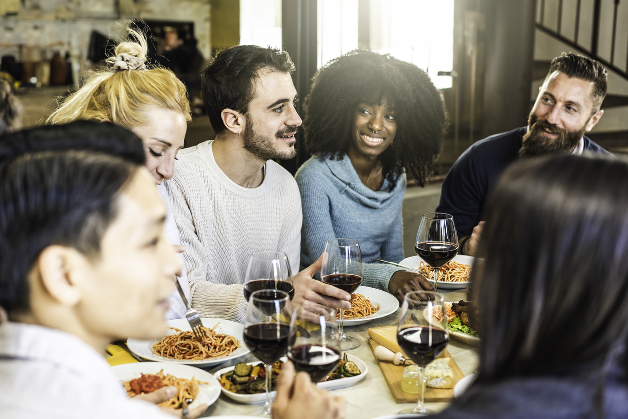A diverse group of friends laughing and talking while eating pasta and drinking red wine at a dinner table.