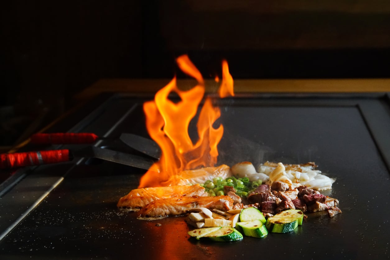 Salmon, seafood, and vegetables cooking on a teppanyaki grill with a large flame rising from the surface.