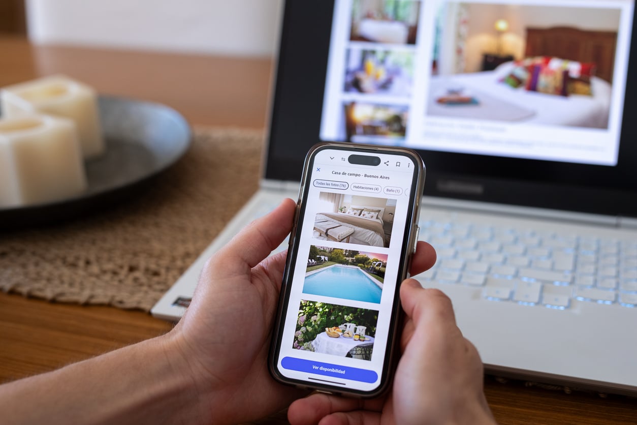 Hands holding a smartphone displaying a travel accommodation listing with photo previews, placed in front of an open laptop.
