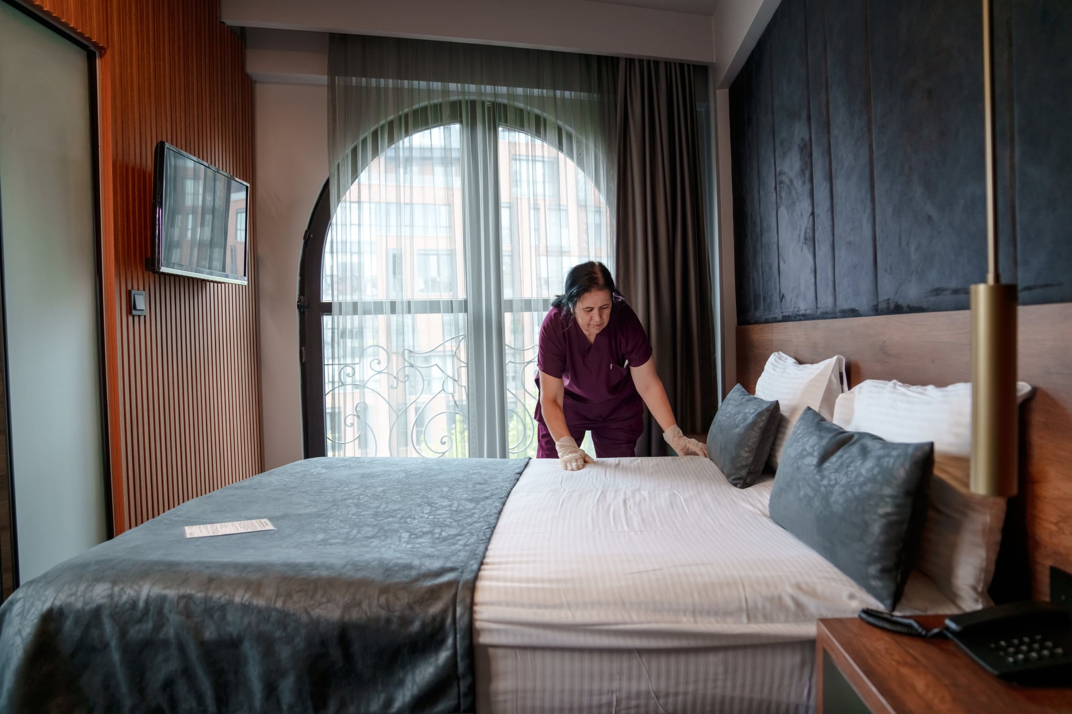 A housekeeper wearing purple scrubs and gloves making a hotel bed.