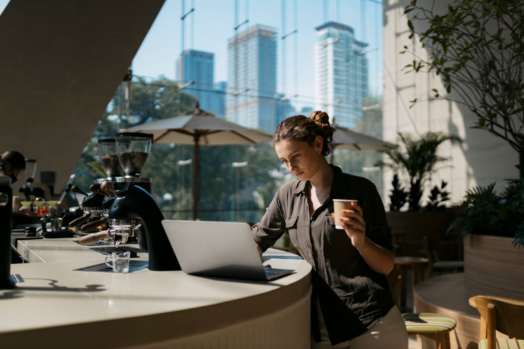 A woman working on a laptop at a coffee shop counter while holding a paper cup.