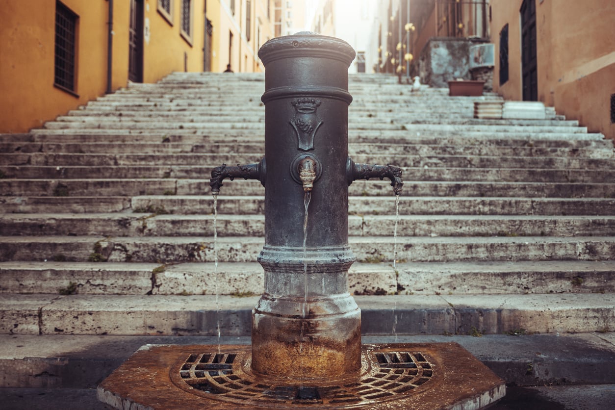 An iron street fountain with three spouts dripping water, situated at the base of a wide stone staircase.