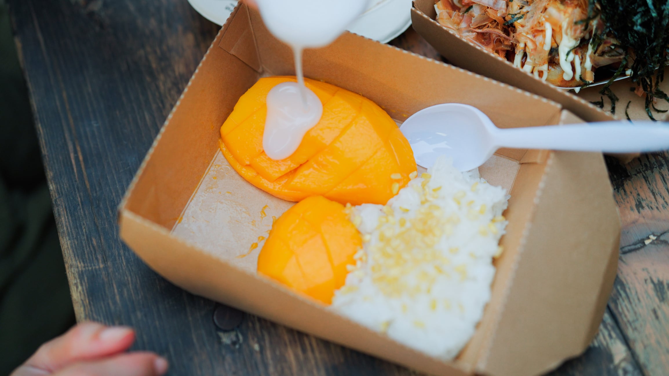 A paper container filled with mango sticky rice being drizzled with coconut milk.