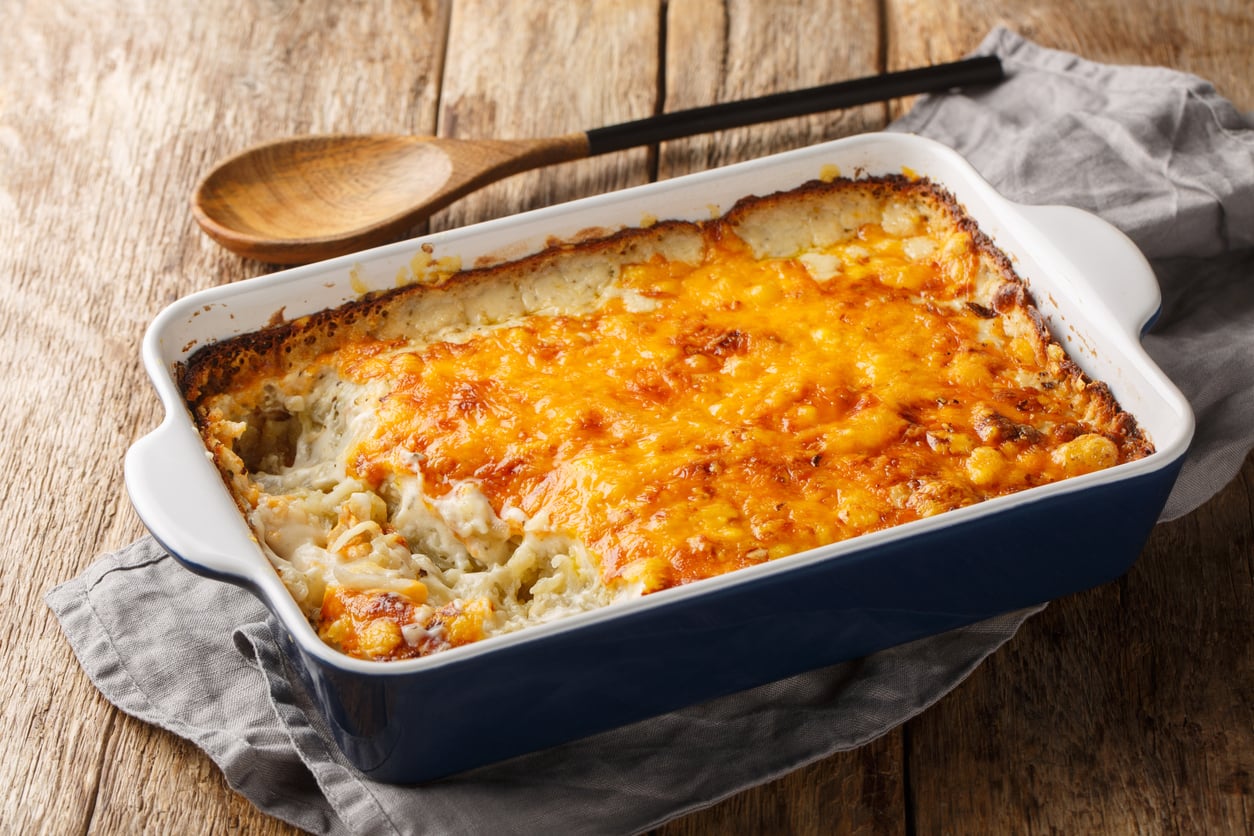 A blue baking dish containing a cheesy pasta bake, with a wooden spoon resting nearby on a cloth-covered wooden surface.