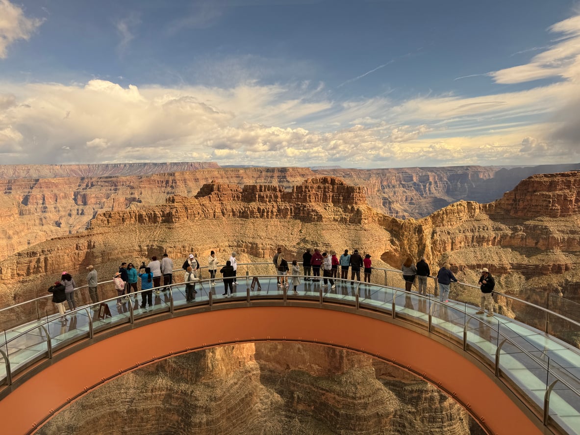 A group of people walk along the U-shaped glass-floored Grand Canyon Skywalk, one of the region’s popular tourist attractions, overlooking a deep desert canyon.