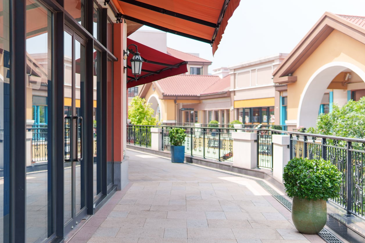 A walkway lined with storefronts, potted plants, and red awnings in an outdoor shopping center.
