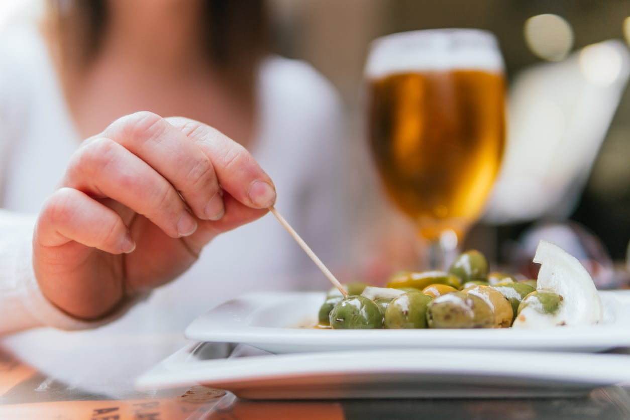 A person picking a green olive from a small plate with a toothpick, with a glass of beer in the background.