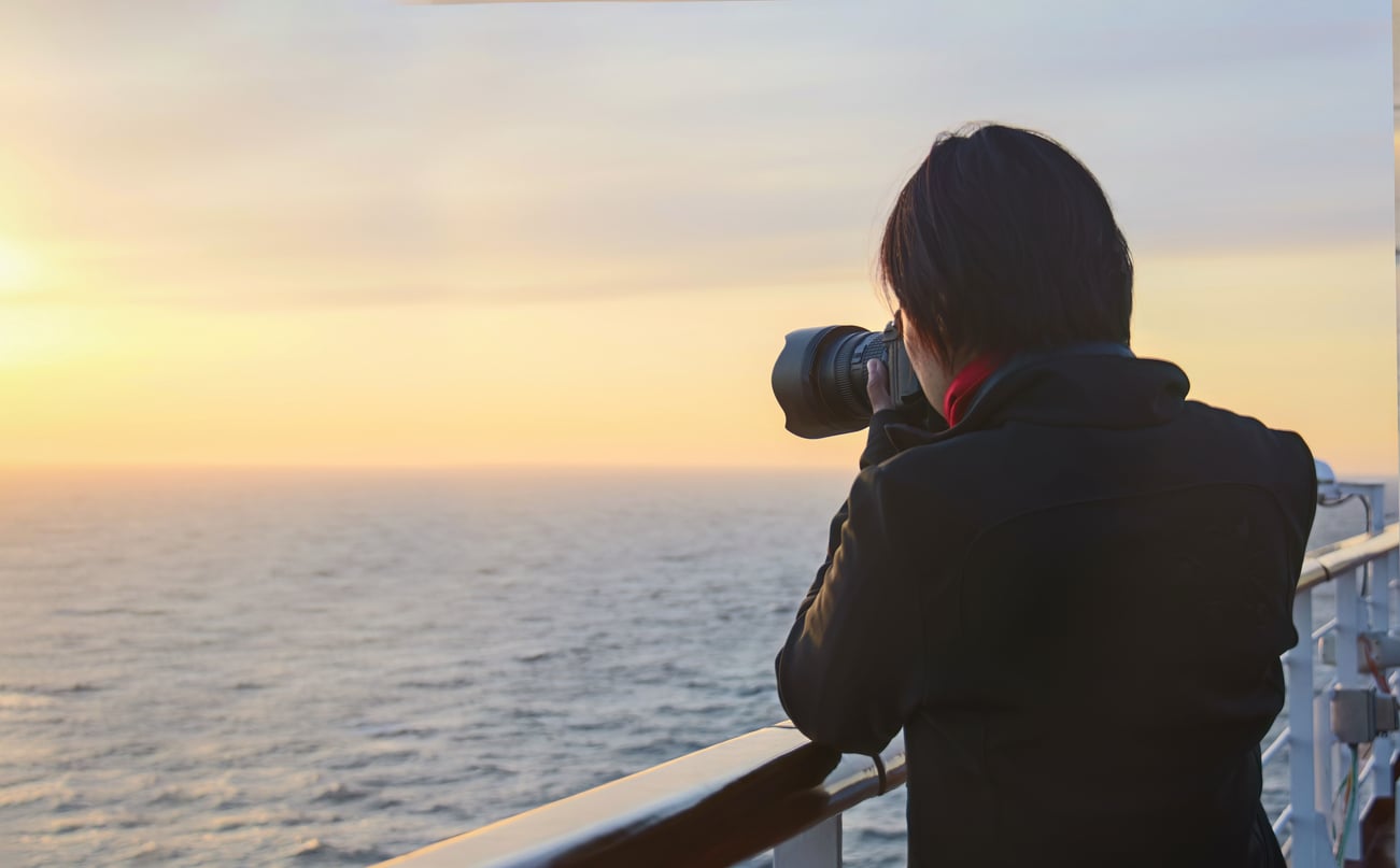 A person stands on the deck of a cruise ship, using a camera to photograph the sunset over the ocean.