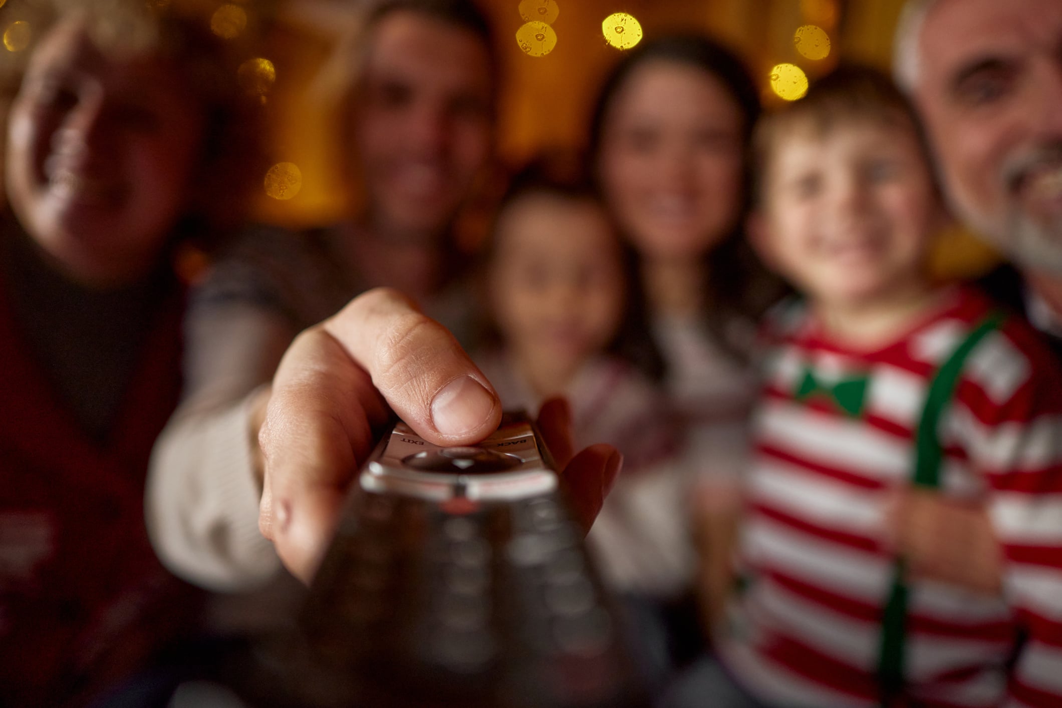 A hand holds a television remote control in the foreground, with a blurred family smiling in the background.