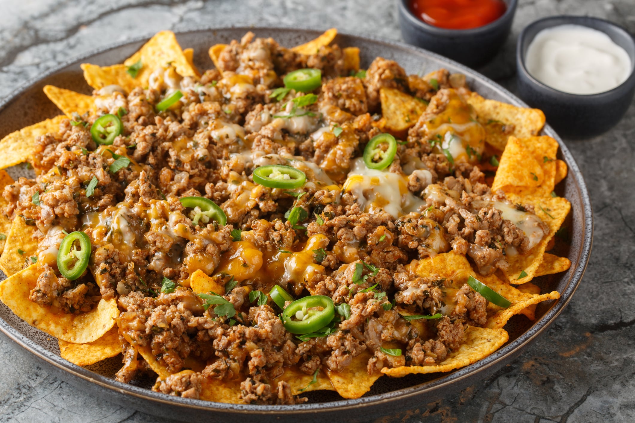 A plate of nachos topped with ground meat, melted cheese, and jalapeño slices, with small bowls of red sauce and sour cream on the side.