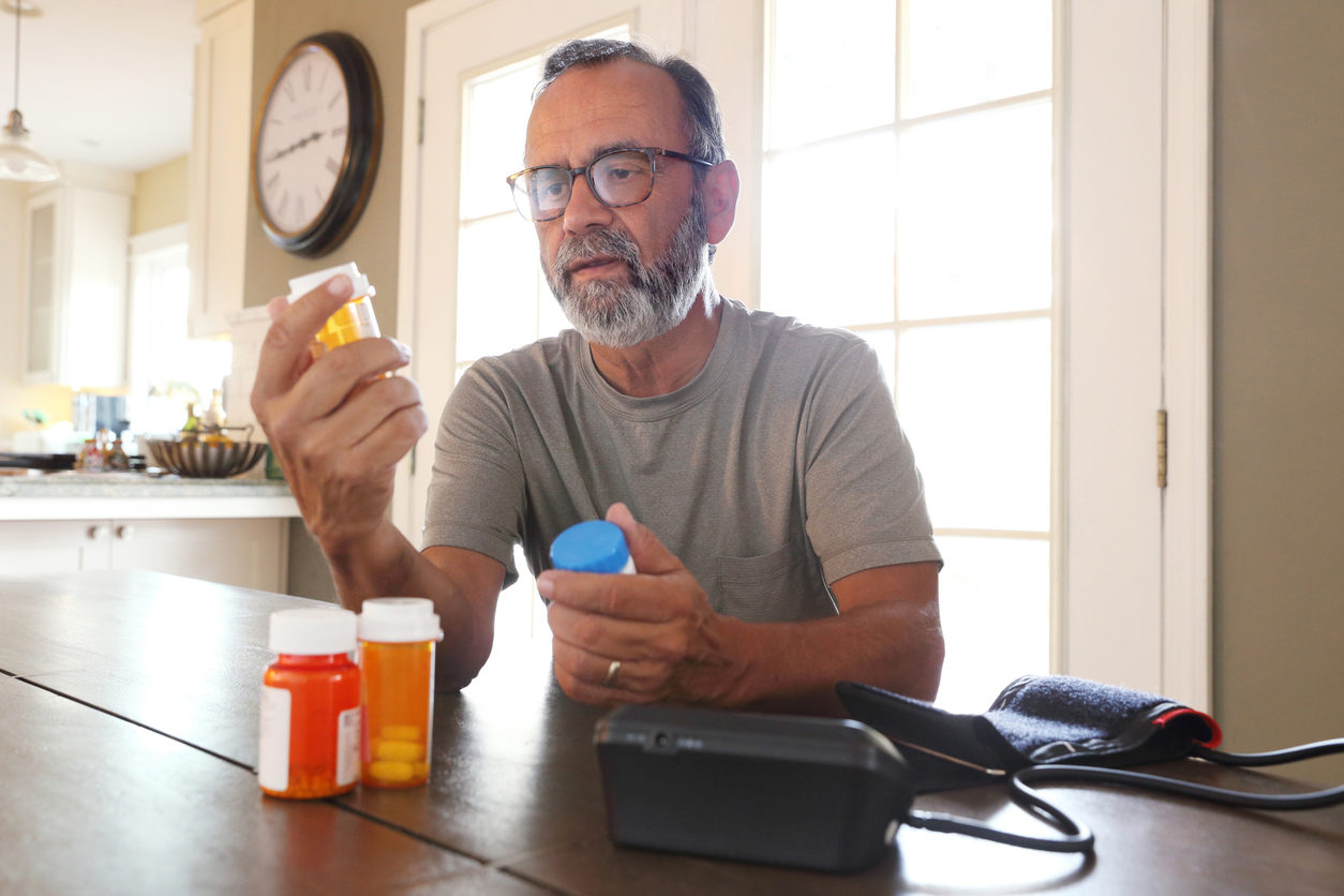 A middle-aged man with a beard and glasses sits at a table reading a prescription medicine bottle, with two other pill bottles and a blood pressure monitor nearby.