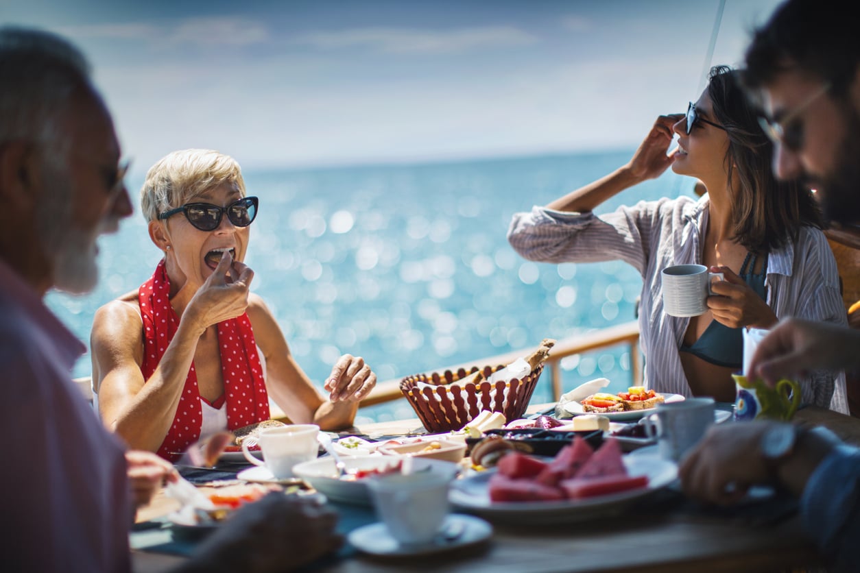 Four people enjoy breakfast at an outdoor table overlooking the ocean during cruise entertainment.
