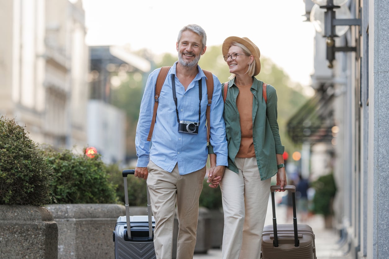 A mature couple holding hands and pulling rolling suitcases while walking down a city sidewalk.