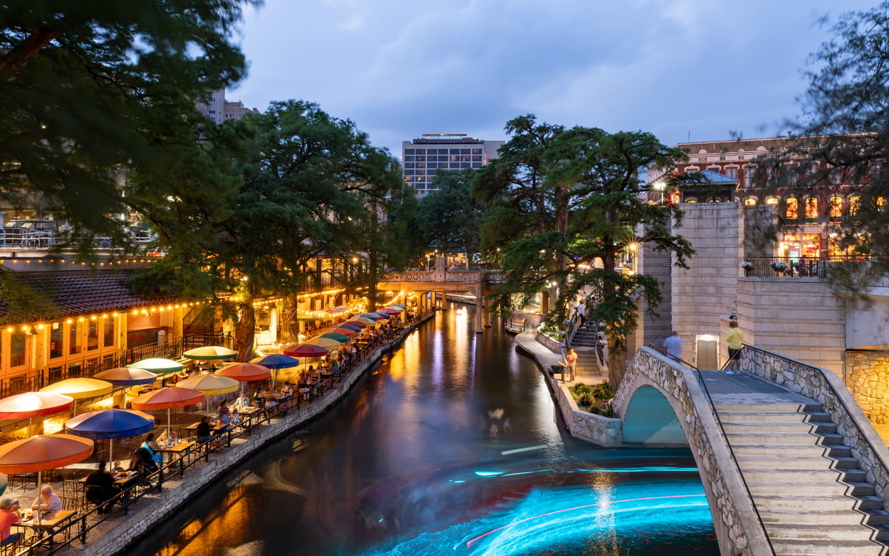 The San Antonio River Walk at dusk with colorful umbrellas shading restaurant patios along the water and a stone footbridge in the foreground.