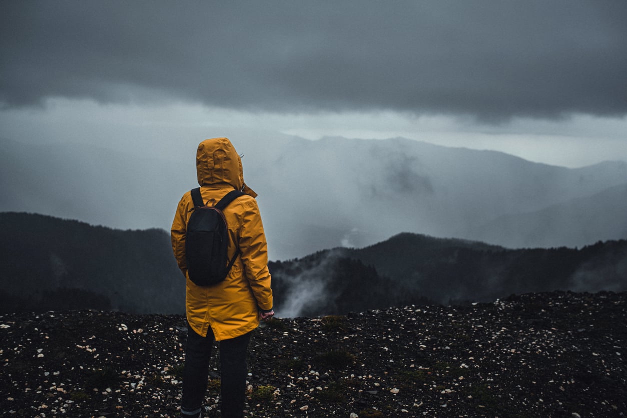 A person in a yellow raincoat and backpack stands on a rocky summit overlooking misty mountains under a stormy sky.