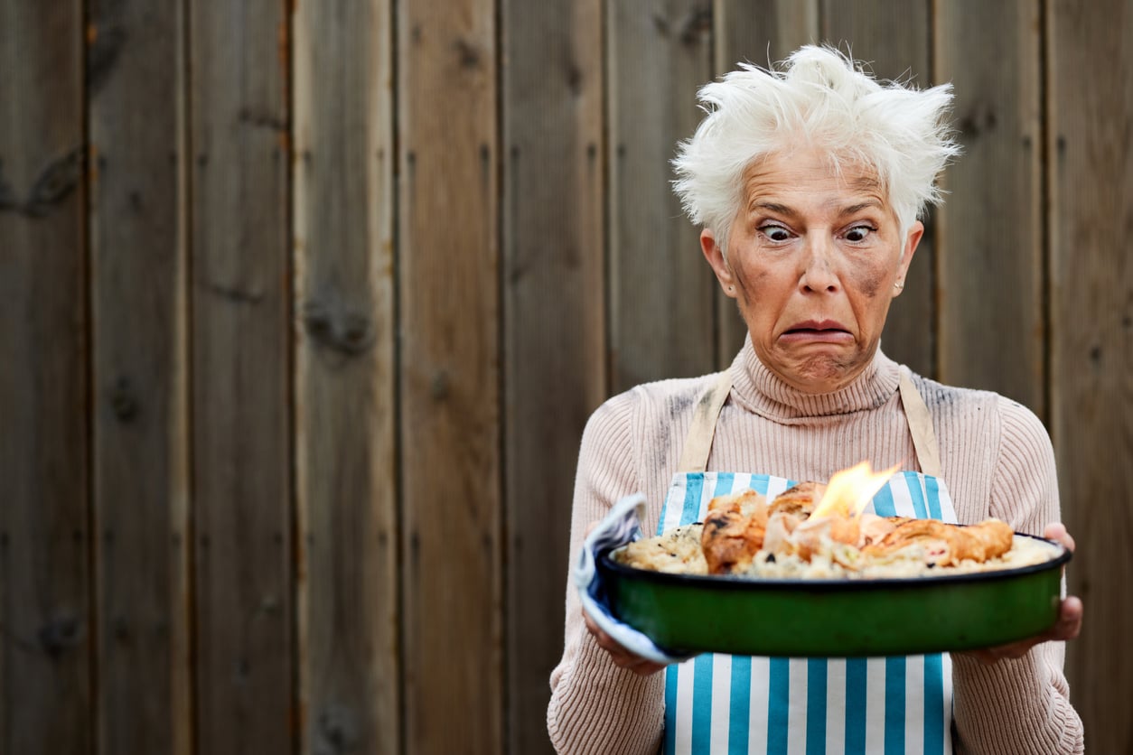 A woman with a messy hairstyle and smudged face looks horrified while holding a pan of food that is on fire.