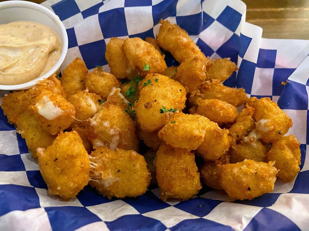 A basket of golden-fried cheese curds in a blue and white checkered liner, served with a side of dipping sauce.