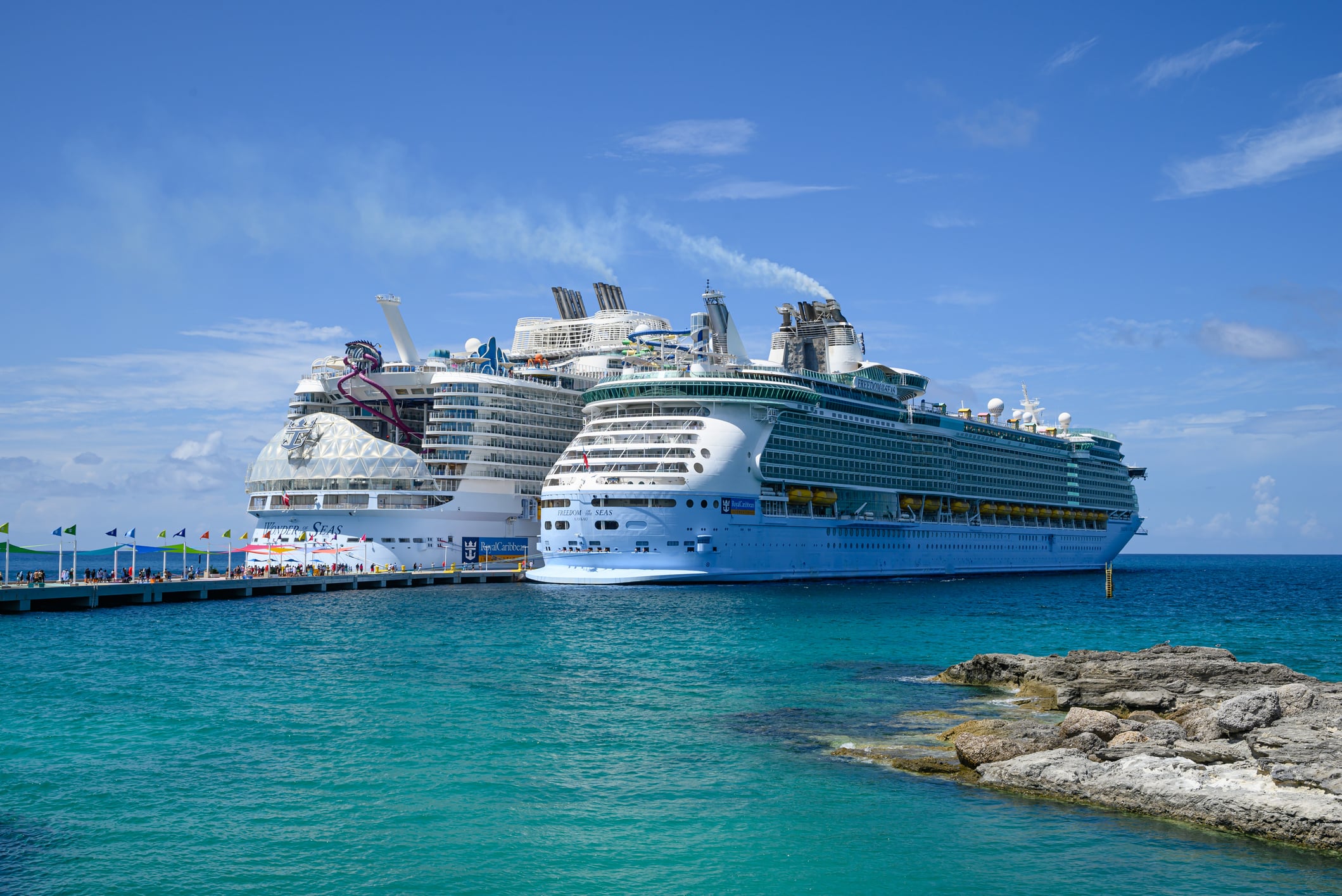 Two large Royal Caribbean cruise ships, Wonder of the Seas and Freedom of the Seas, docked at a pier in turquoise water.