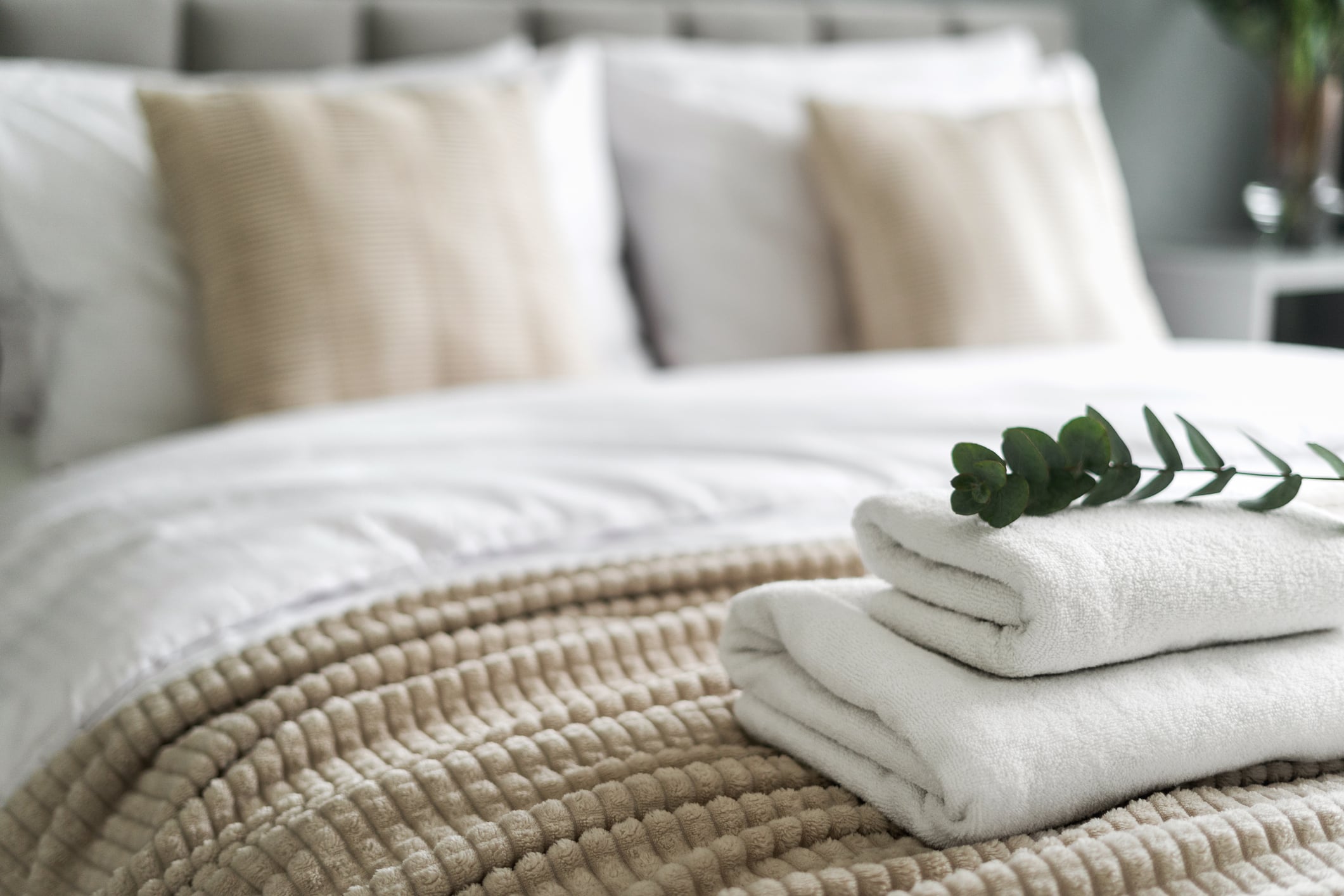 Two folded white towels topped with a sprig of eucalyptus on a bed with a textured tan throw blanket.