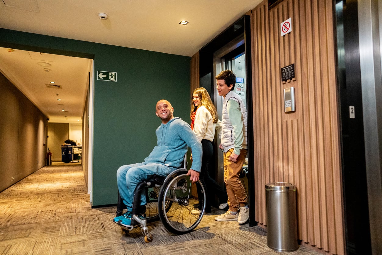 A man in a wheelchair and two teenagers exit a hotel elevator into a hallway, ready to begin their trip planning decisions.