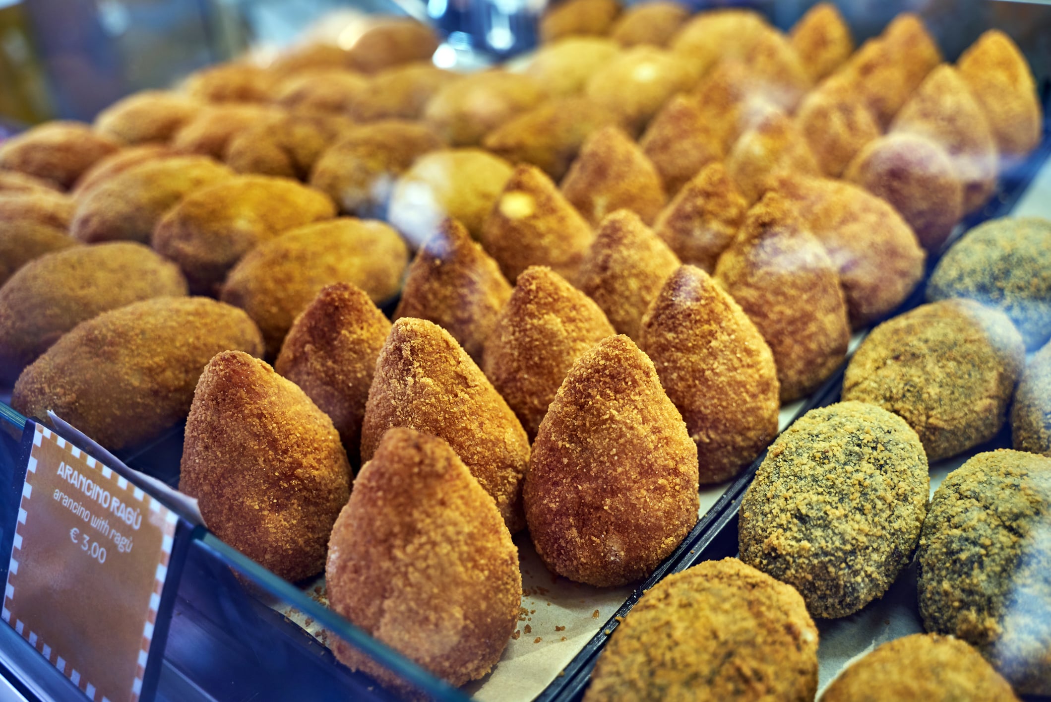 Rows of fried, cone-shaped arancini and rounded snacks displayed in a glass deli case with a sign for “Arancino Ragù” priced at €3.00.