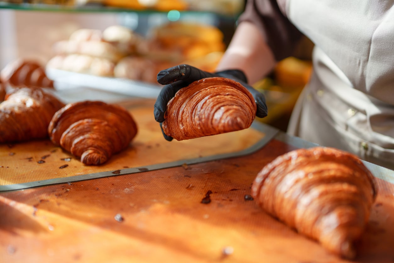 A baker wearing a black glove picks up a freshly baked croissant from a tray.