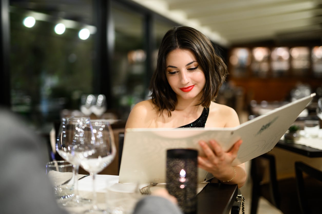 A woman with red lipstick reviews a restaurant menu at a table set with a candle and wine glasses.