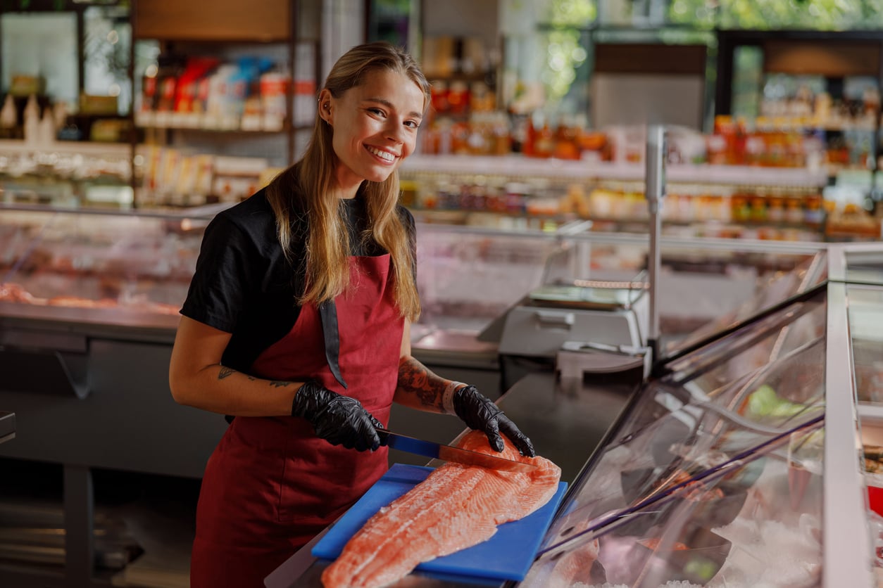 Smiling employee in a red apron and black gloves cutting a salmon fillet on a blue cutting board behind a deli counter.